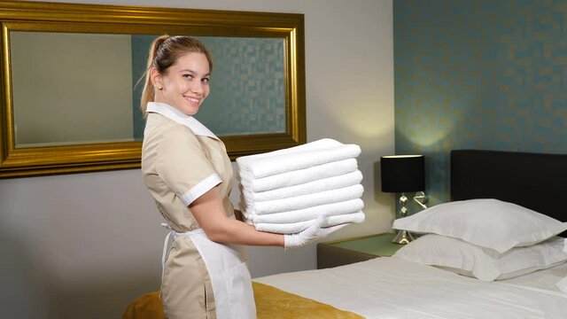 Professional cleaning service. Pretty maid holding stack of folded clean towels then throwing it up smiling at camera with king-sized bed in background. Room preparation for guests. 4 k video