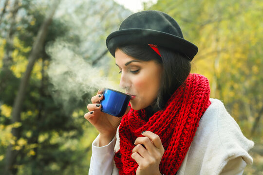 A woman in a white coat, crochet Red scarf and black hat holds a cup in her hands. Tea party in nature