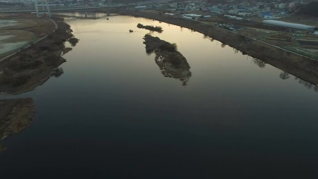 Aerial footage flying over and vacant field and river with a small island in the middle toward a bridge and small community in background just before sunset