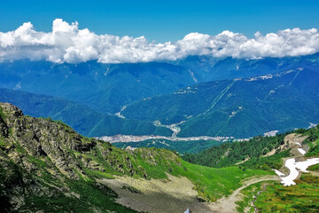 Obraz premium Alpine landscape. In the valley there is green grass, islands of snow. A village is visible below. The peaks of the mountain range are hidden in the clouds. Summer. Caucasus. Russia.
