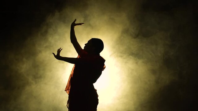 Silhouette a young woman dancer in a red sari. Indian folk dance. Shot in a dark studio with smoke and neon lighting.