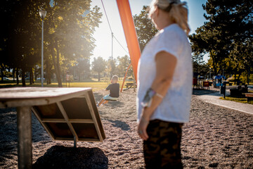 Young boy playing on a zip line in public park during summer day. He is with grandmother.