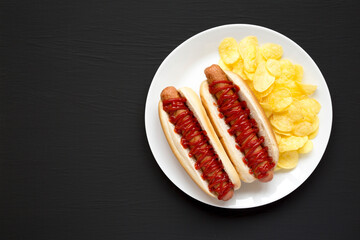 Tasty American Hot Dog with Potato Chips on a white plate on a black surface, top view. Flat lay, overhead, from above. Copy space.