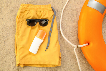 Male swimsuit with sunglasses, sunscreen and rescue ring on sand