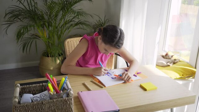 concentrated brunette teenager girl in pink blouse draws in paper notebook with marker sitting at wooden table near window in light room on sunny day