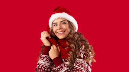 Portrait of sweet young lady wearing woolen sweater, Santa hat and scarf on red background