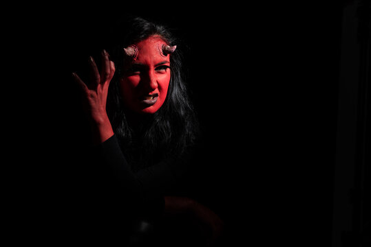 Halloween concept. Headshot of angry woman with red face makeup, wears black clothes, wants to look spooky, poses against black background with copy space. Demon woman with red face and small horns
