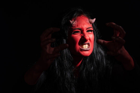 Halloween concept. Headshot of angry woman with red face makeup, wears black clothes, wants to look spooky, poses against black background with copy space. Demon woman with red face and small horns