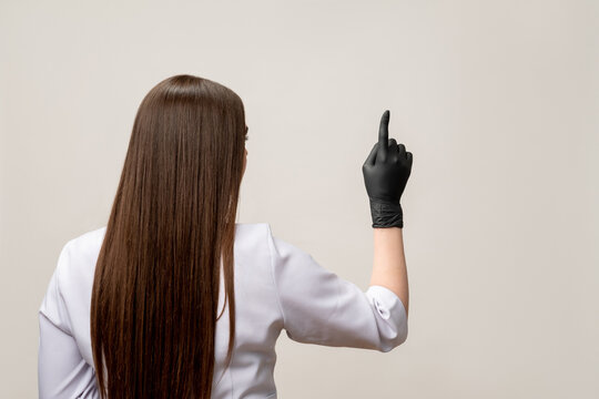 Beauty procedure. Master class. Brunette woman in white medical overall black latex gloves pointing at copy space isolated on light background. Professional microblading. Rejuvenation cosmetology.