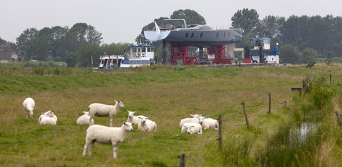 Transport of a super yacht on a pontoon with tugboats at the river. . Ship building industry. Super sailing yacht. Netherlands. Zwarte Water river. Dike with sheep. Dutch landscape. Genemuiden.