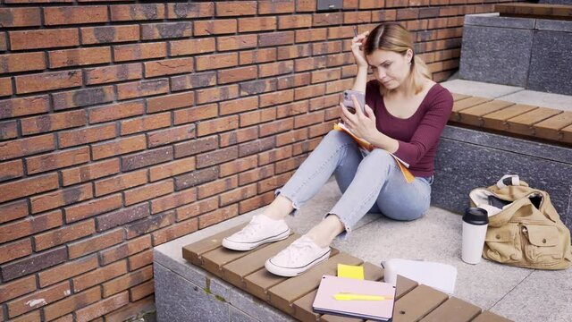 young stressed woman student scores out something in paper notebook with pen looking at smartphone screen on stone bench on modern city street