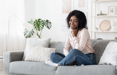 Obraz premium Shy Black Girl Sitting On Sofa At Home, Posing To Camera
