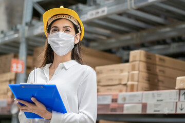Female construction worker wearing protective face mask writing on clipboard checking product on aisle in warehouse factory. industrial social responsibility prevention Coronavirus. new normal working