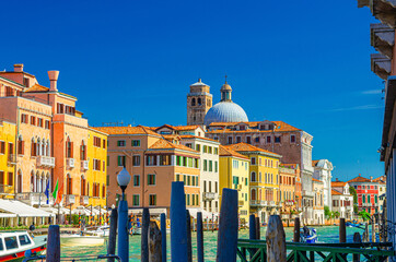 Venice Grand Canal waterway with old colorful buildings, tower and dome of Chiesa di San Geremia church, pier with wooden poles in water, blue sky background in summer day, Veneto Region, Italy