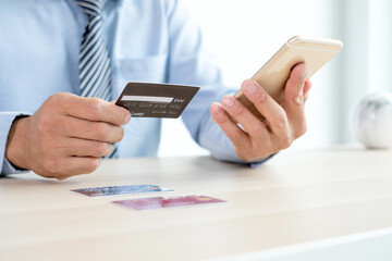 The businessman's hand is holding a credit card and using a smartphone for online shopping and internet payment in the office