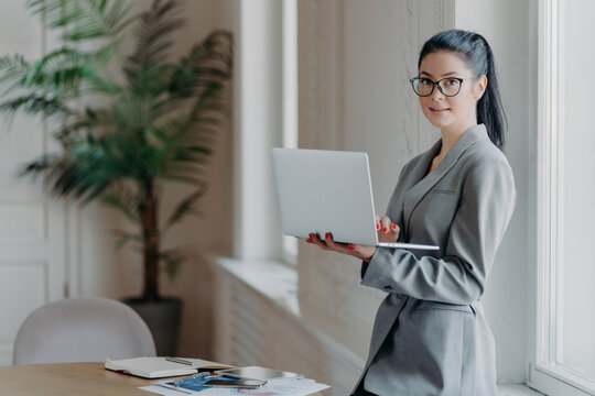 Woman Administrative Stands Near Desktop, Holds Modern Laptop Computer, Uses Wireless Connection To Internet, Dressed In Formal Grey Outfit, Searches Information On Web Page, Works Distantly