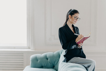 Attracative successful businesswoman in elegant clothes records information in notepad, sits on back of sofa, poses in home office, plans working process, makes list to do, writes down points