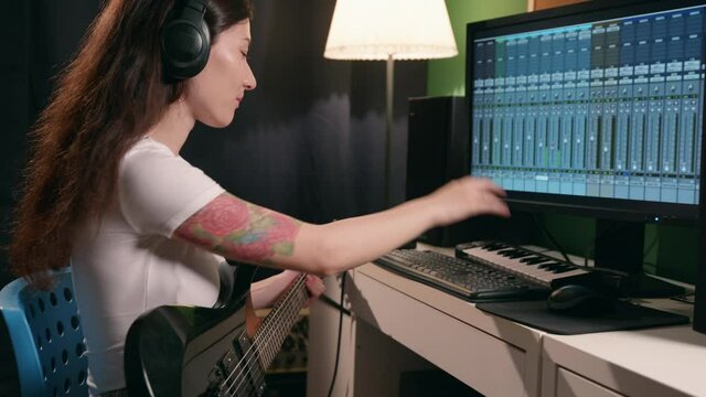 Girl enjoying process of creativity. Female musician recording song in home record studio in soft cinematic light. Young woman playing guitar and synthesizer creating music using equalizer program.