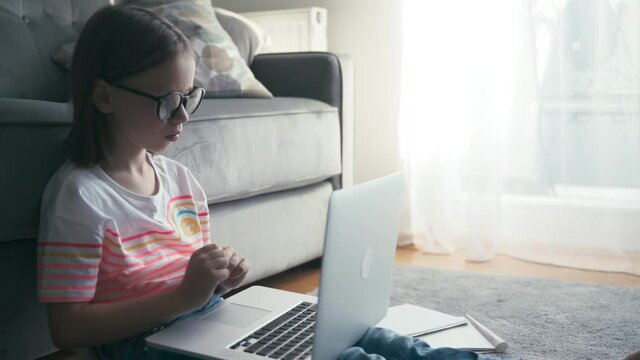 Serious little 8 years old girl in glasses sits on a carpet in a living room typing on a laptop and making notes in a workbook. Remote education technologies and homework.