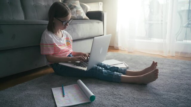 Serious little 8 years old girl in glasses sits on a carpet in a living room typing on a laptop and making notes in a workbook. Remote education technologies and homework.