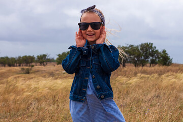 Adorable little girl in denim jacket, black sunglasses, blue plaid dress in yellow grass field. Happy stylish long blonde hair child on countryside landscape. Cute kid walking outdoor rural road trip.