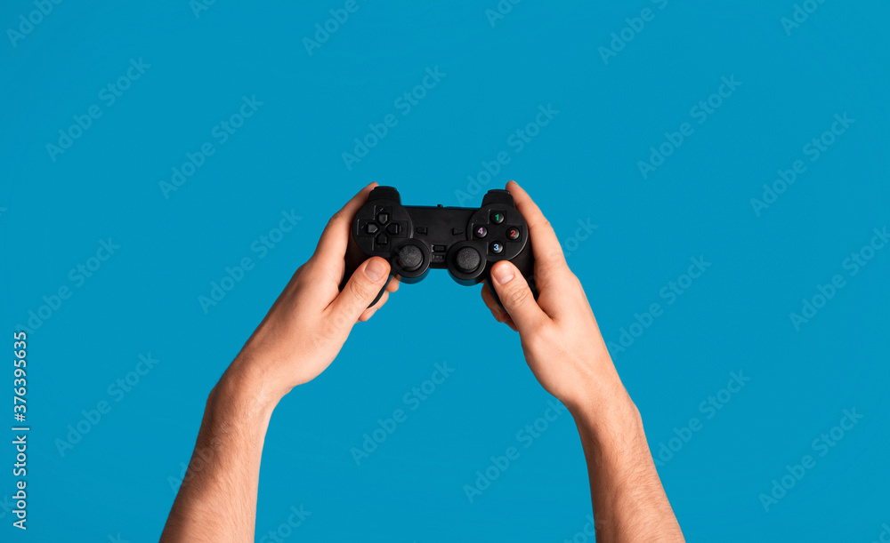 Wall mural young man holding videogame joystick on blue background, closeup of hands