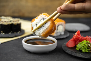 take a philadelphia maki rolls with bamboo chopsticks and put in into  soy sauce. pink ginger, wasabi on black stone plate, asian food, japanese cuisine background
