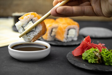 take a philadelphia maki rolls with bamboo chopsticks and put in into  soy sauce. pink ginger, wasabi on black stone plate, asian food, japanese cuisine background
