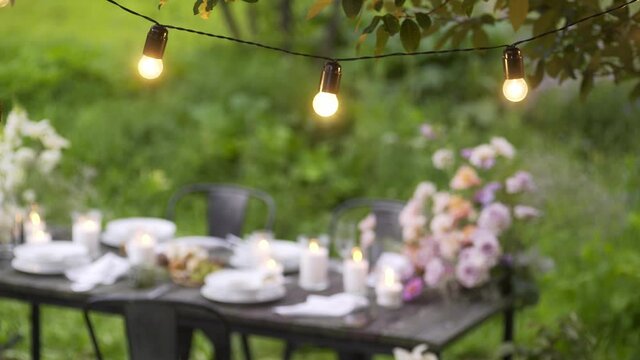 vintage glowing fairy lights hang on tree branches against blurred view of festive served table with burning candles and fresh flowers closeup