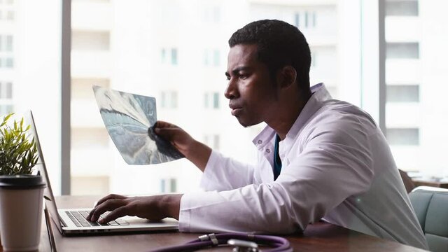 Side view of African American black male doctor in white coat analyzing history disease of patient using MRI brain head scan, working on laptop, sitting at desk in light office on background of window