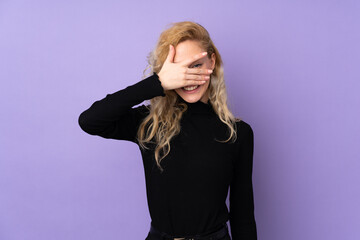 Young blonde woman isolated on purple background covering eyes by hands and smiling