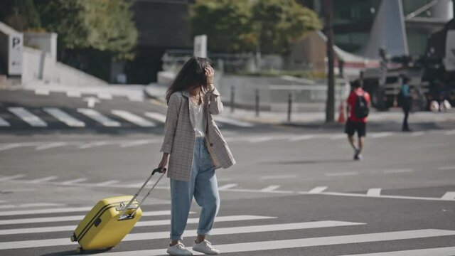 Portrait of young woman walkinig across the street towards camera, rolling suitcase behind. Girl walking on crosswalk with luggage. Background of easy wide crossroad. Concept of travel, lifestyle.