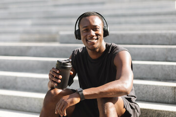 Positive Black Sportsman In Wireless Headphones Resting After Jogging Outdoors, Drinking Water