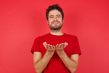 Young handsome caucasian man wearing t-shirt over isolated red background holding something with open palms, offering to the camera.