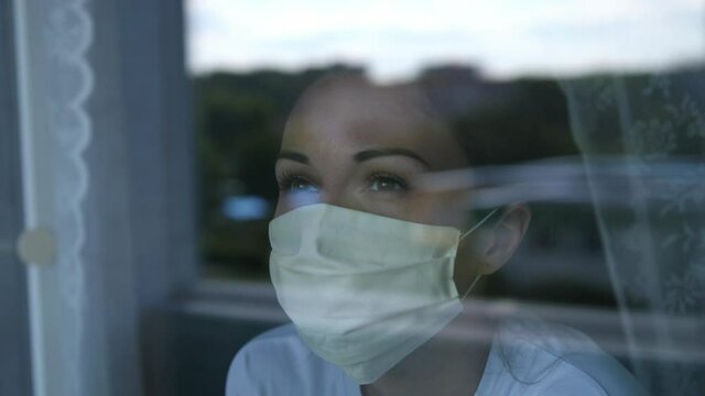Young woman wears a mask to protect herself from covid-19 while looking out the window. slow motion