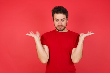 Hesitant Young handsome caucasian man wearing t-shirt over isolated red background shrugs shoulders, looks uncertain and confused. Have no answer