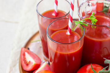 Fresh red tomato juice in a glass with a straw and jar with tomatoes on light wooden background.