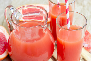 Fresh red grapefruit juice in a glass with a straw and jar with fruit pieces on light wooden background.