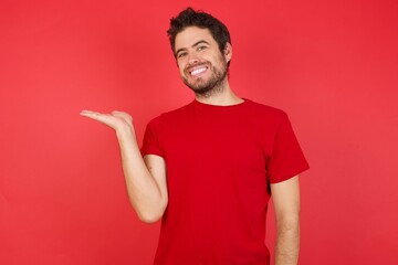 Young handsome caucasian man wearing t-shirt over isolated red background smiling cheerful presenting and pointing with palm of hand looking at the camera.