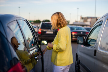 A young woman with a coronavirus mask in a yellow sweater gets into her car. The car is parked in the parking. There is bokeh in the background. Coronavirus concept. Life with covid-19. 