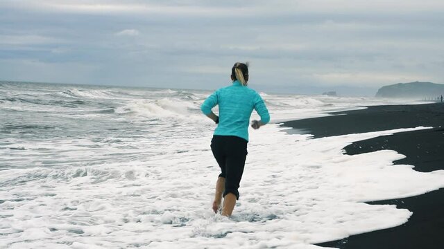 Young active woman athlete running on black volcanic beach exercising female runner training at pacific ocean background. Concept healthy jogging and outdoor exercise