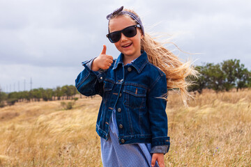 Adorable little girl in denim jacket, black sunglasses, blue plaid dress in yellow grass field. Happy stylish long blonde hair child on countryside landscape. Cute kid walking outdoor rural road trip.