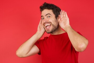 Naklejka premium Young handsome caucasian man wearing t-shirt over isolated red background Trying to hear both hands on ear gesture, curious for gossip. Hearing problem, deaf