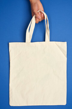 female hand holds an empty beige textile bag by the handles on a blue background