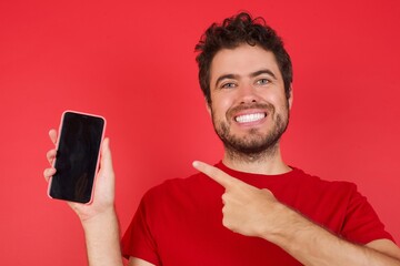 Young handsome caucasian man wearing t-shirt over isolated red background with telephone in hand new model of apple telephone just have look at price