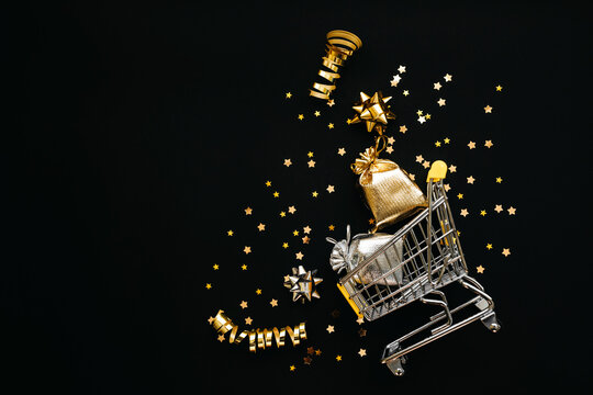 Shopping trolley and various golden items on a black or dark background. Festive conceptual Christmas or New Year background or Black Friday sale.