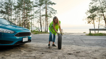 Full length shot of attractive young woman wearing reflective vest, rolling spare tire to change the flat one on the road side