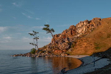 Obraz premium Red stones rock in the light of the setting sun, by the bay with the coast on lake Baikal, reflection, coniferous larch trees