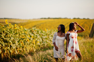 Two pretty young black friends woman wear summer dress pose in a sunflower field.