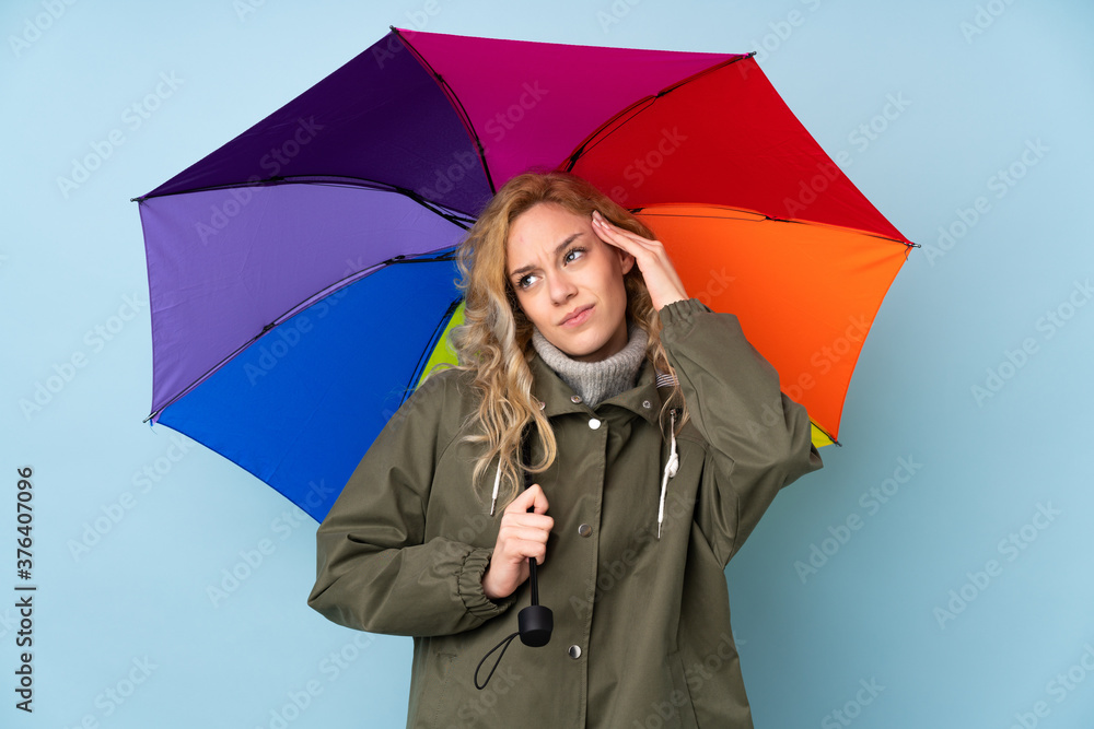 Canvas Prints Young blonde woman holding an umbrella isolated on blue background having doubts and with confuse face expression